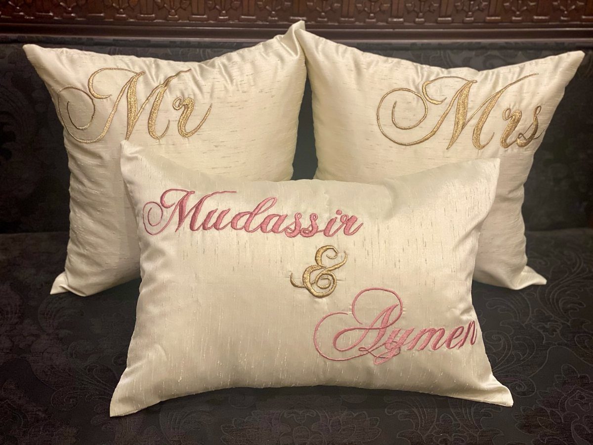Three cream satin pillows with gold and pink embroidery on a dark sofa