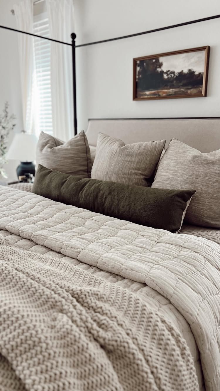 Layered neutral bedding with a quilted coverlet and olive green accent pillow.