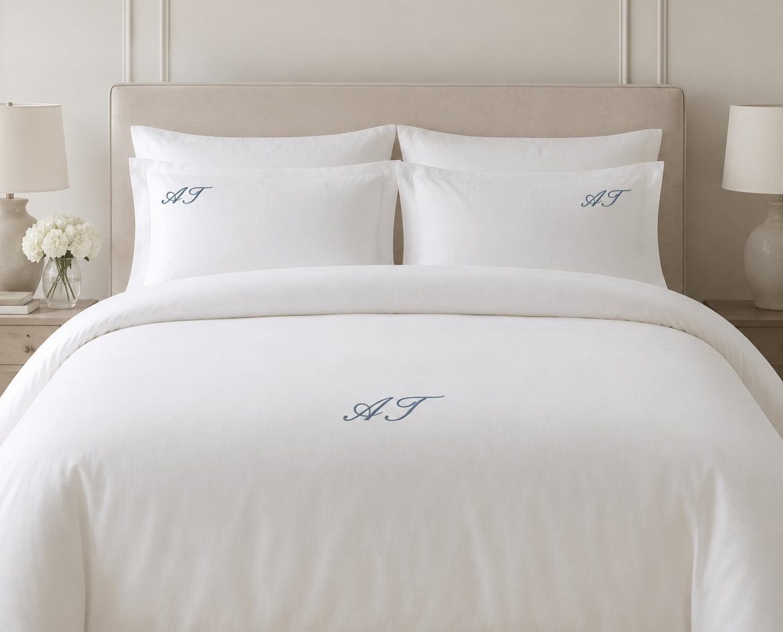 White bedding with blue embroidered initials on a bed with beige headboard.