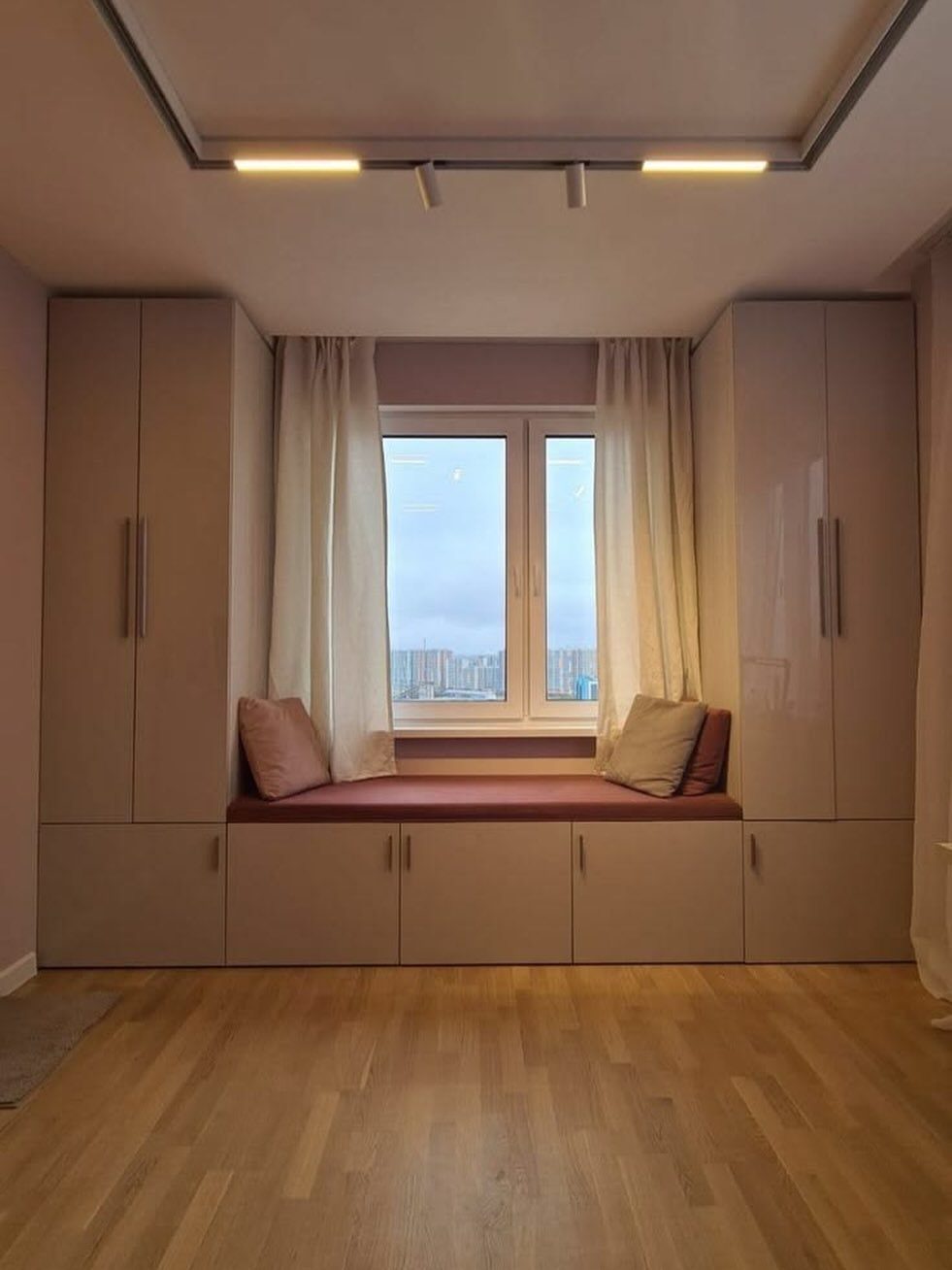 Built-in window seat with maroon cushion between two tall beige wardrobes.