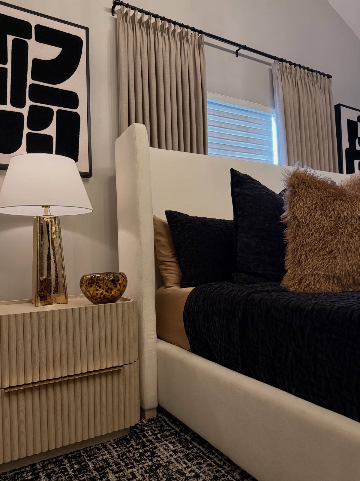 Bedroom with white bed, black duvet, gold fluffy pillow, and gold lamp.