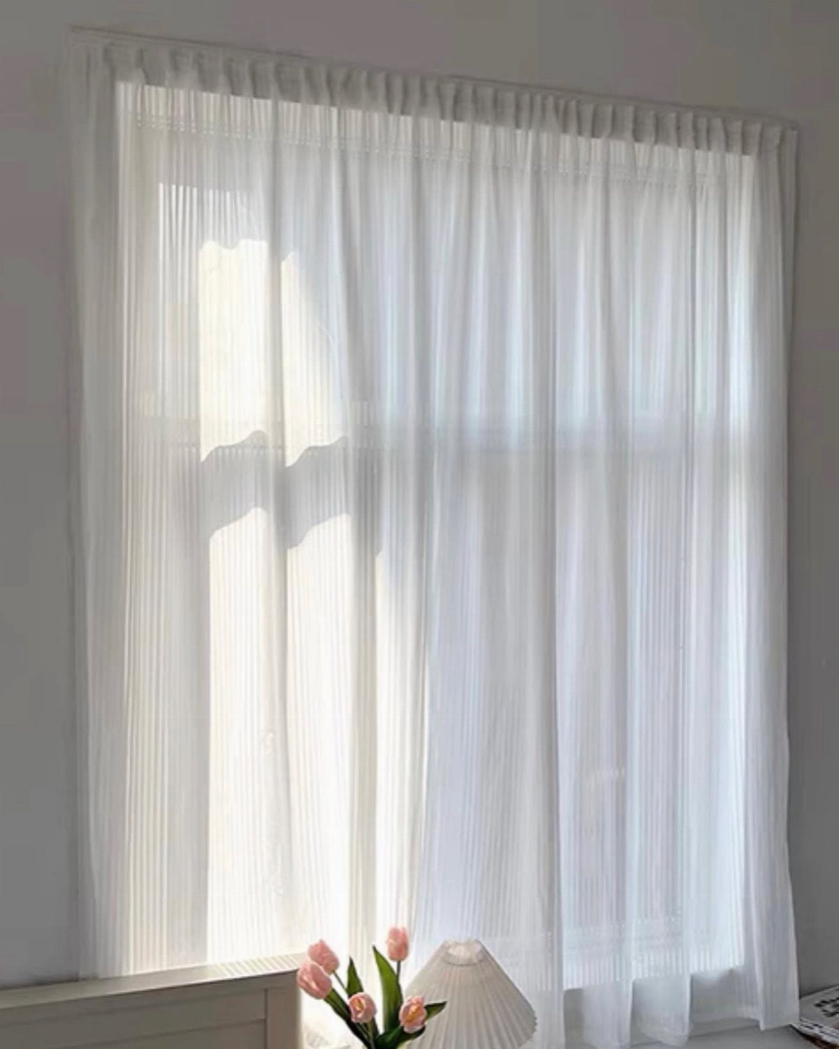 Sheer white curtains on a window against solid white walls with pink tulips.