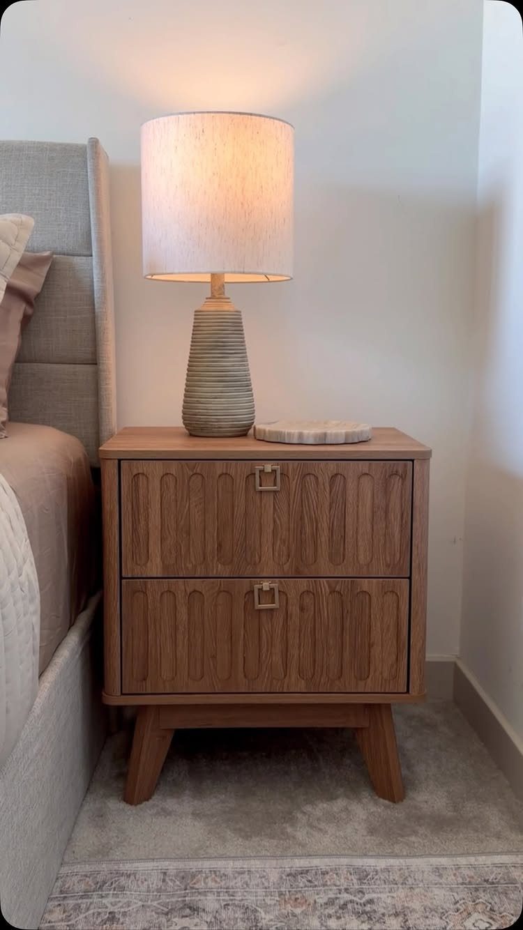 Wooden nightstand with a lit lamp next to a grey upholstered bed