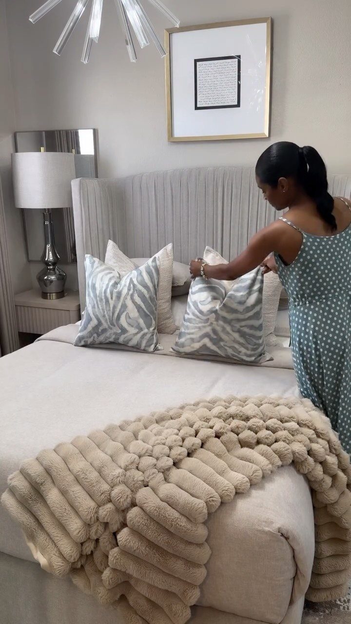 Woman arranging patterned throw pillows on a bed with a chunky knit blanket.