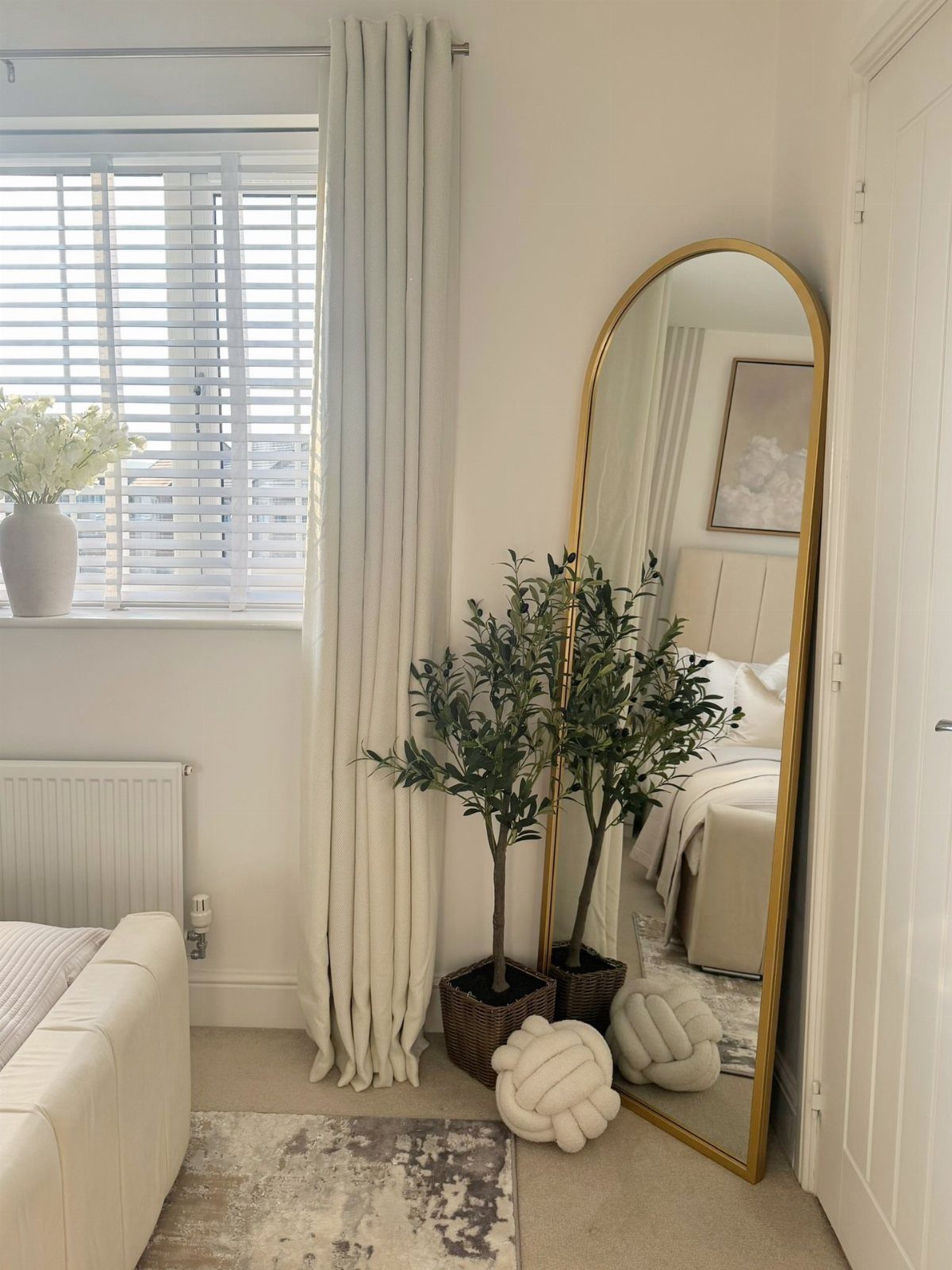 Arched gold-framed mirror leaning against a white wall next to a potted olive tree.