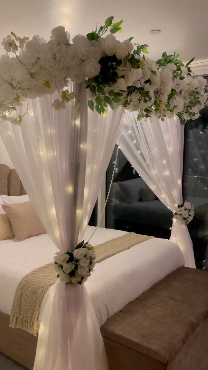 White canopy bed decorated with white roses, greenery, and warm fairy lights.