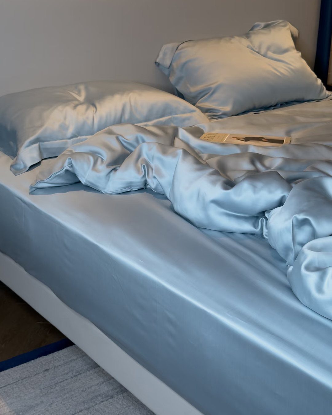 Light blue silk bedding and pillows on a white bed with a magazine.
