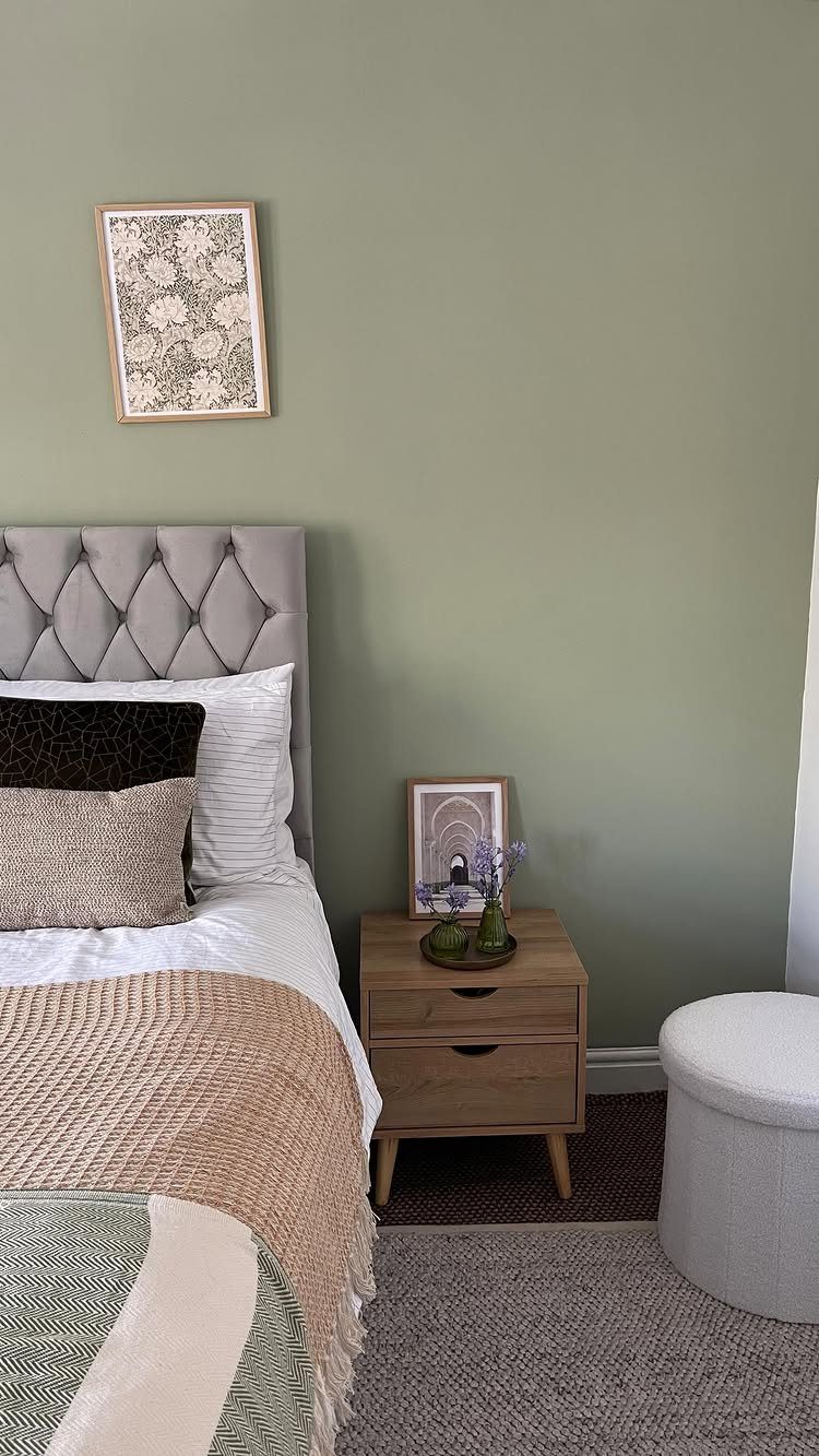 Sage green bedroom wall with a grey tufted headboard and wooden nightstand.