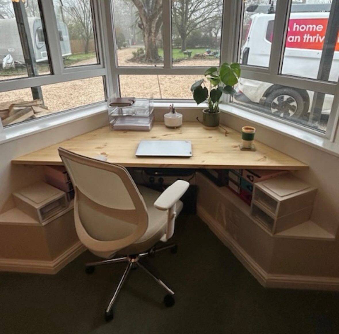 Wooden desk and white office chair built into a bright bay window nook.