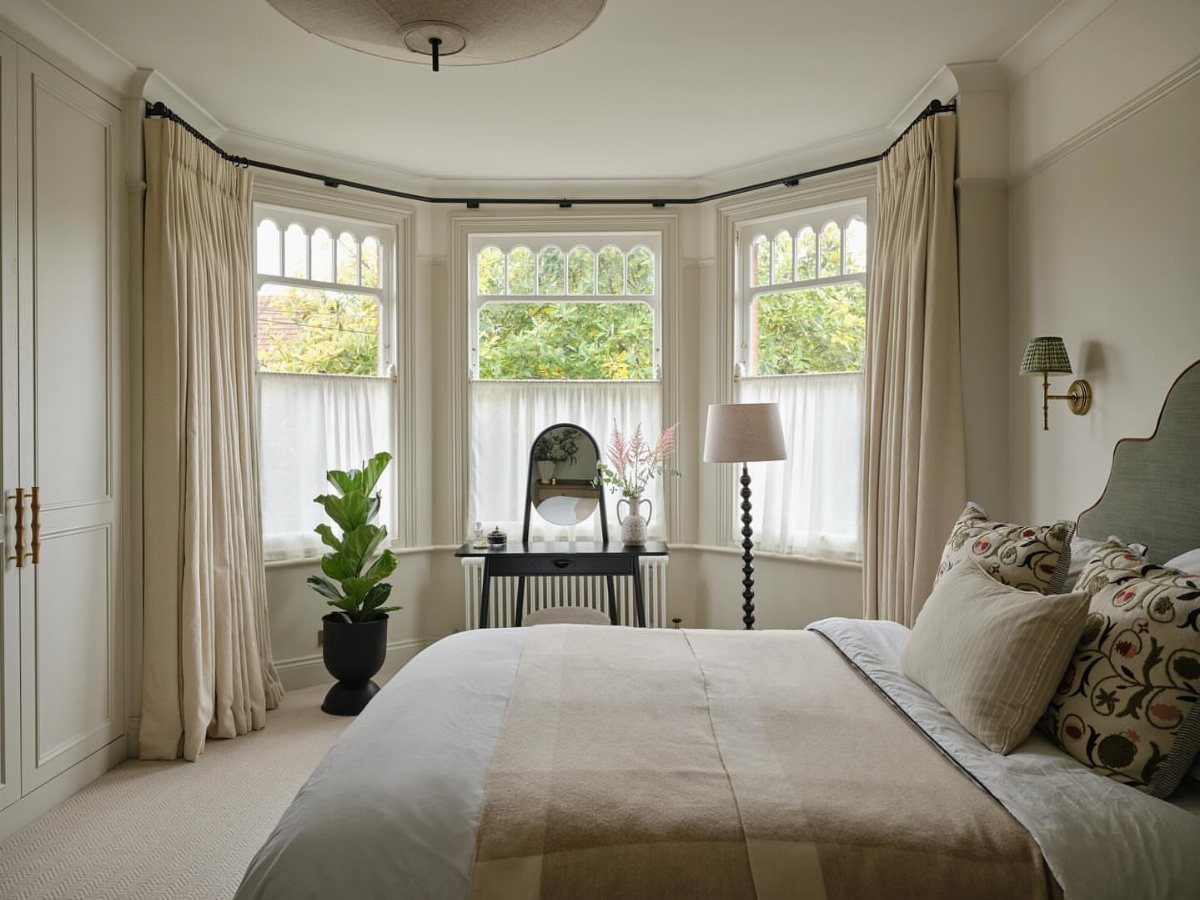 Bedroom with a large bay window, neutral bedding, and cream curtains.