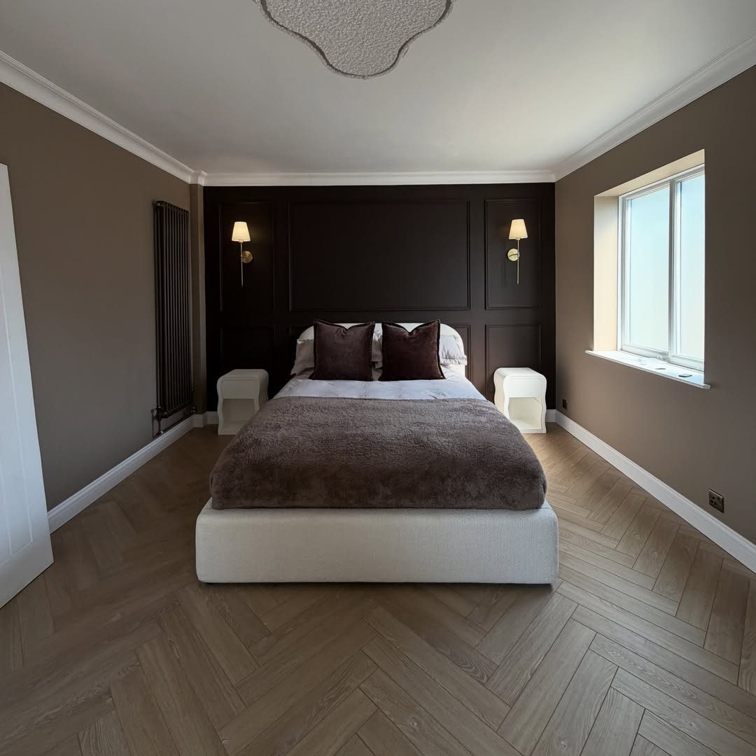 Modern bedroom with light brown herringbone wooden flooring and a white bed.