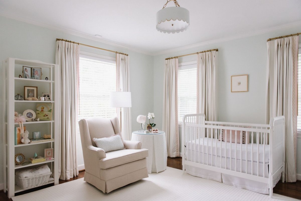 Pale blue-green nursery with a white crib, beige glider, and cream curtains.