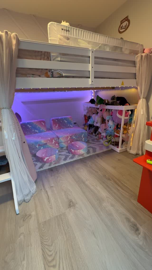 White bunk bed fort with sheer curtains, purple lighting, and colorful floor cushions.
