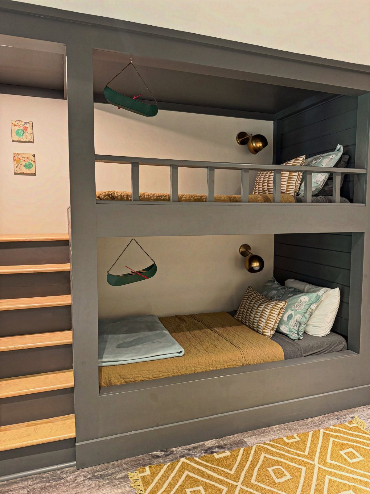 Built-in grey bunk beds recessed into a wall with wooden stairs and gold lamps.