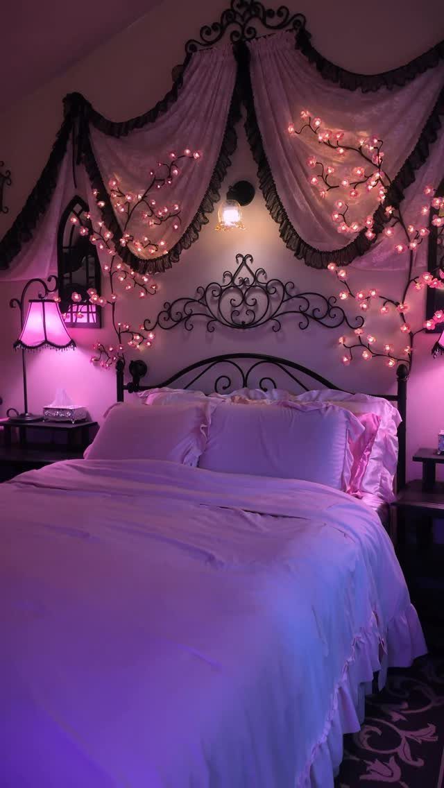 Bed with purple linens and ornate headboard under draped fabric and pink fairy lights.