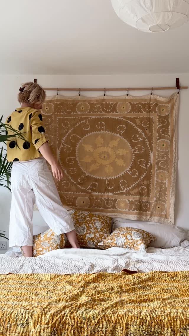 Person adjusting a large patterned tan tapestry hanging above a bed.