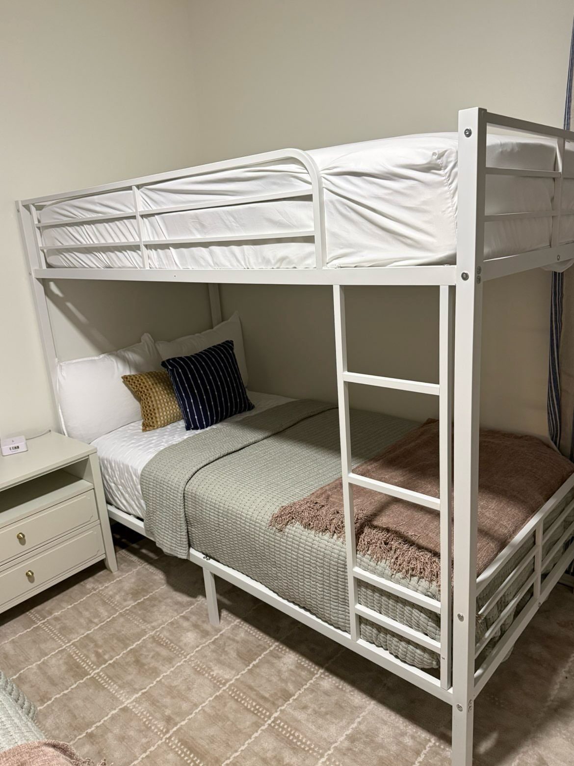 White metal bunk bed with neutral bedding and a small white nightstand.