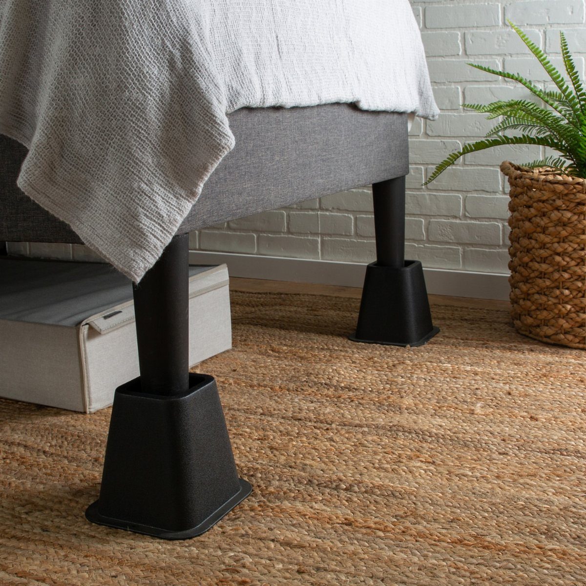 Black bed risers lifting a bed frame above a brown woven jute rug.