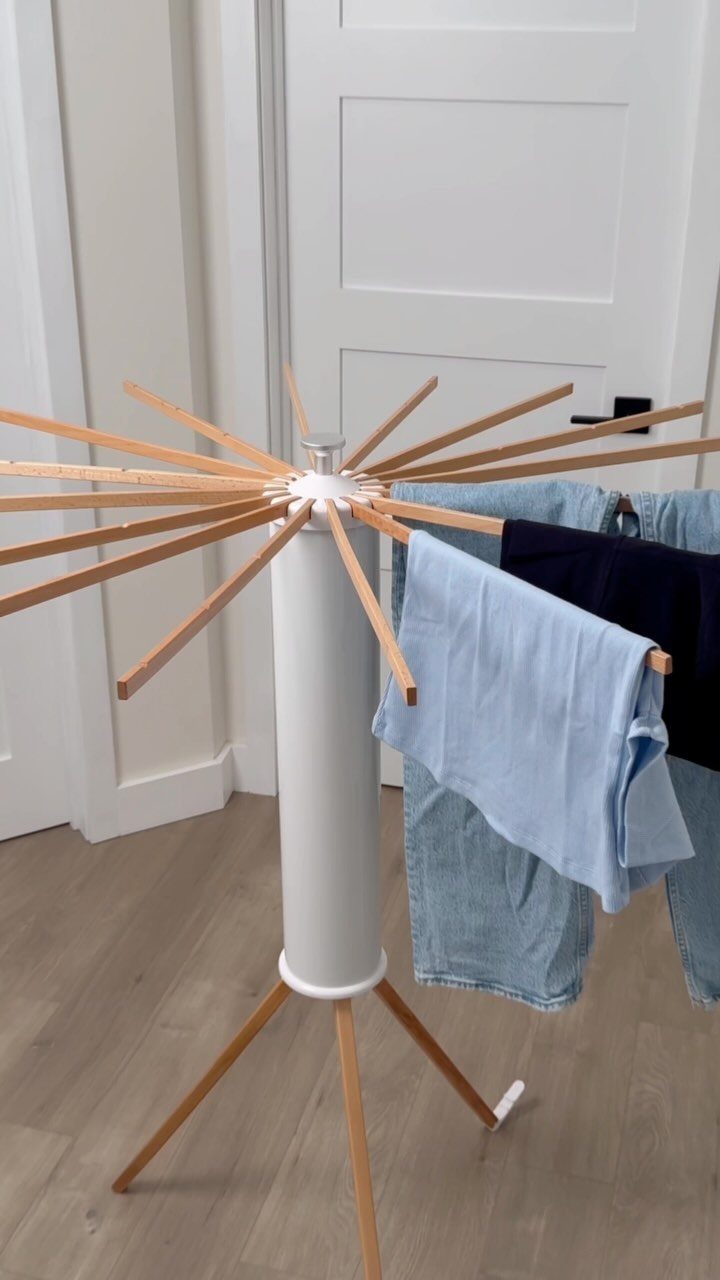 White cylindrical drying rack with wooden arms holding blue and dark clothing indoors.