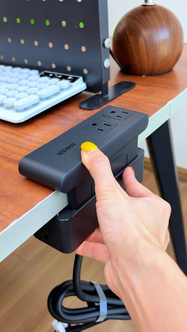 Black Anker power strip clamped to a wooden desk being touched by a hand.