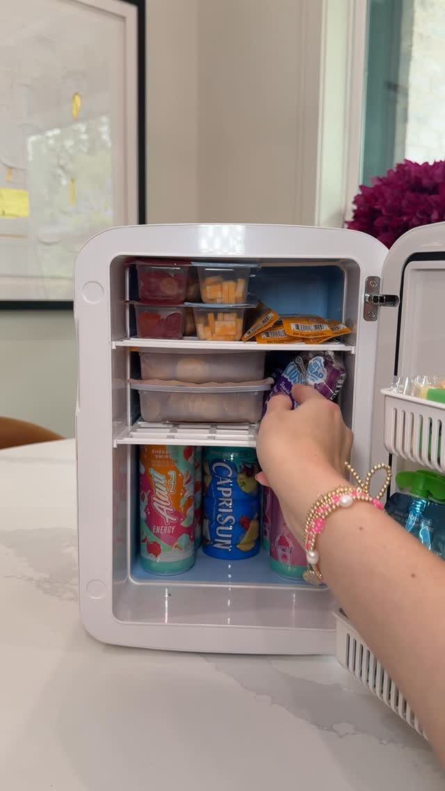 Small white mini fridge filled with snacks and drinks on a white counter.