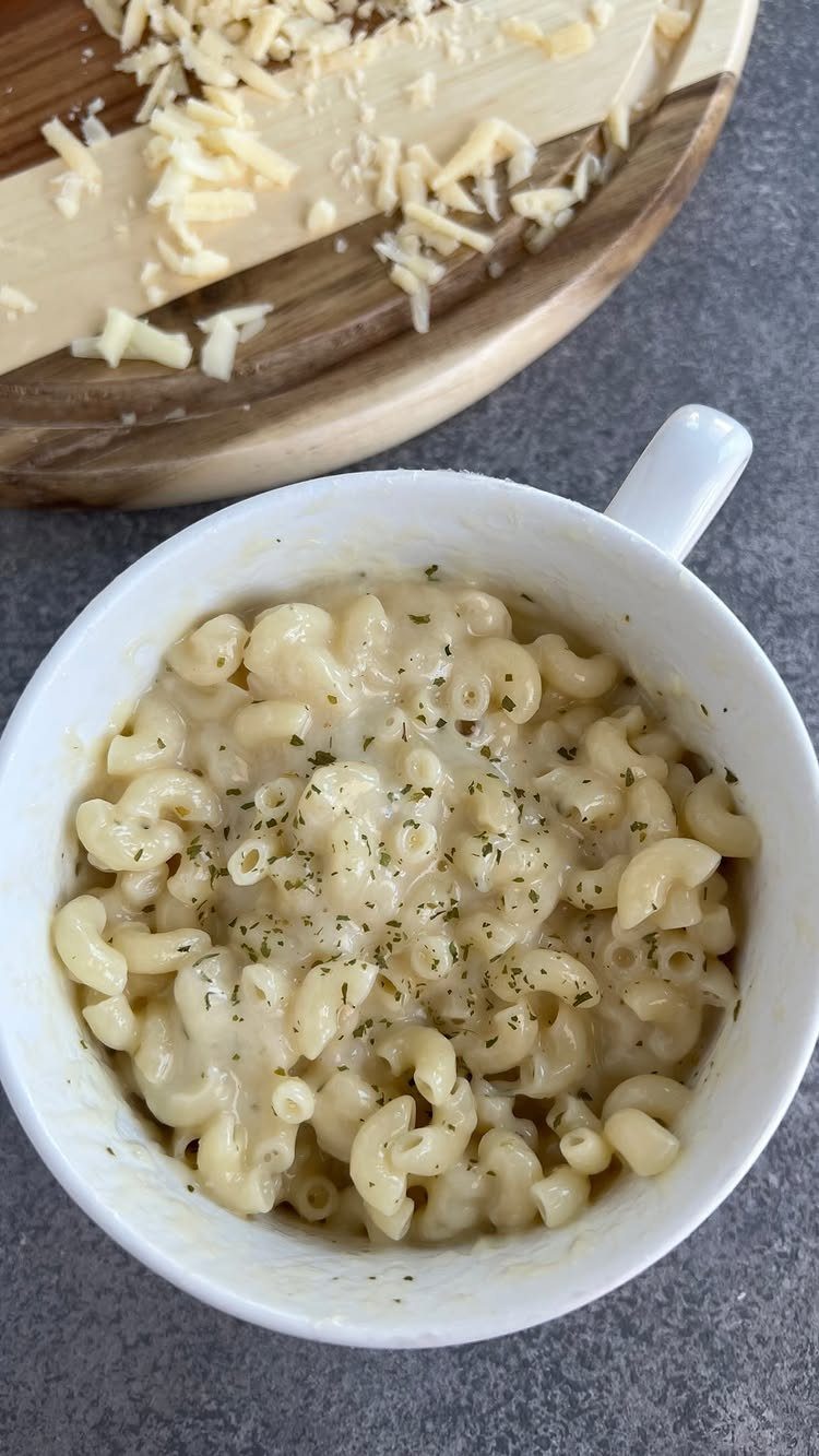 Macaroni and cheese in a white mug topped with herbs and shredded cheese.
