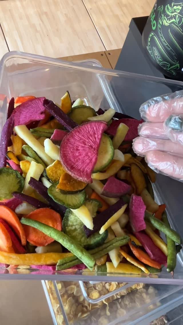 Colorful dried vegetable chips in a clear plastic storage container on a wooden table.