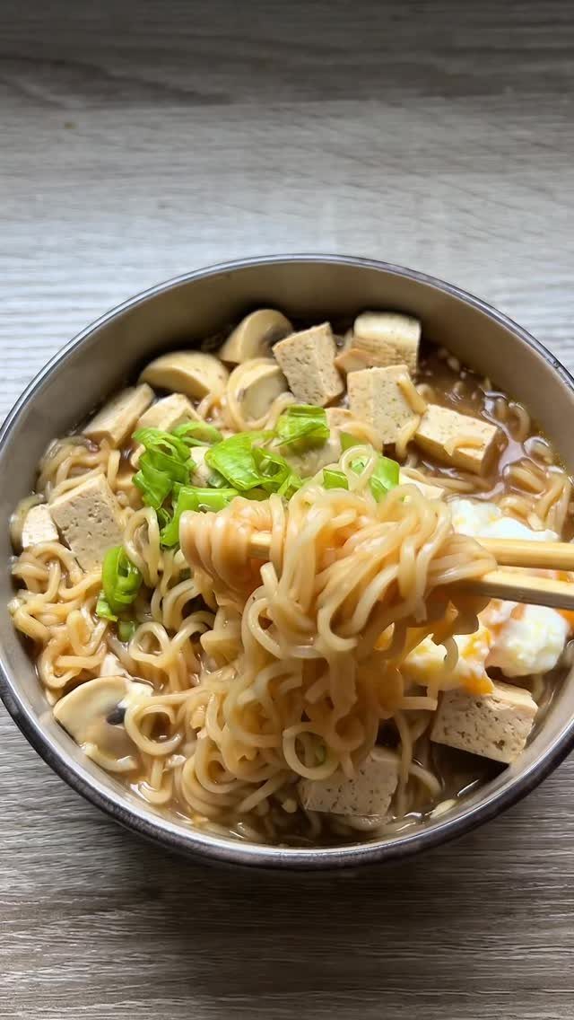 Ramen noodles with tofu, mushrooms, and green onions lifted by wooden chopsticks.