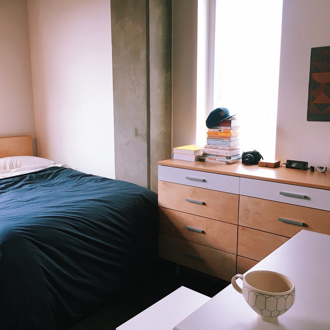 Small bedroom with blue bedding, wooden dresser, and books by a bright window.