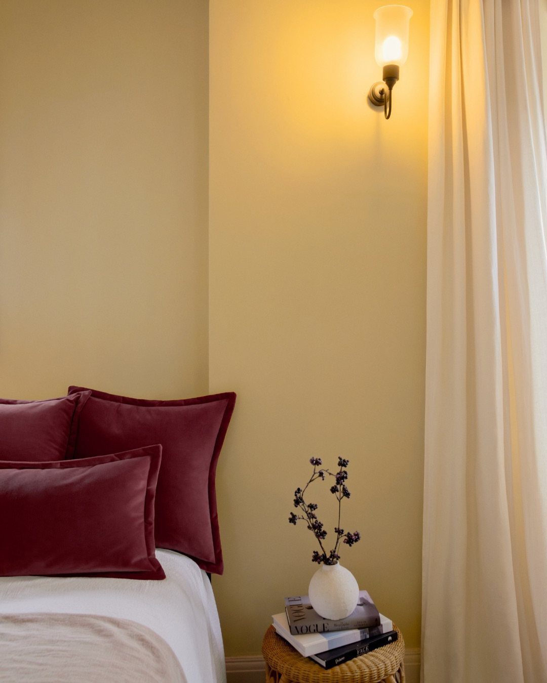 Pale yellow bedroom wall with red velvet pillows and a glowing wall sconce.