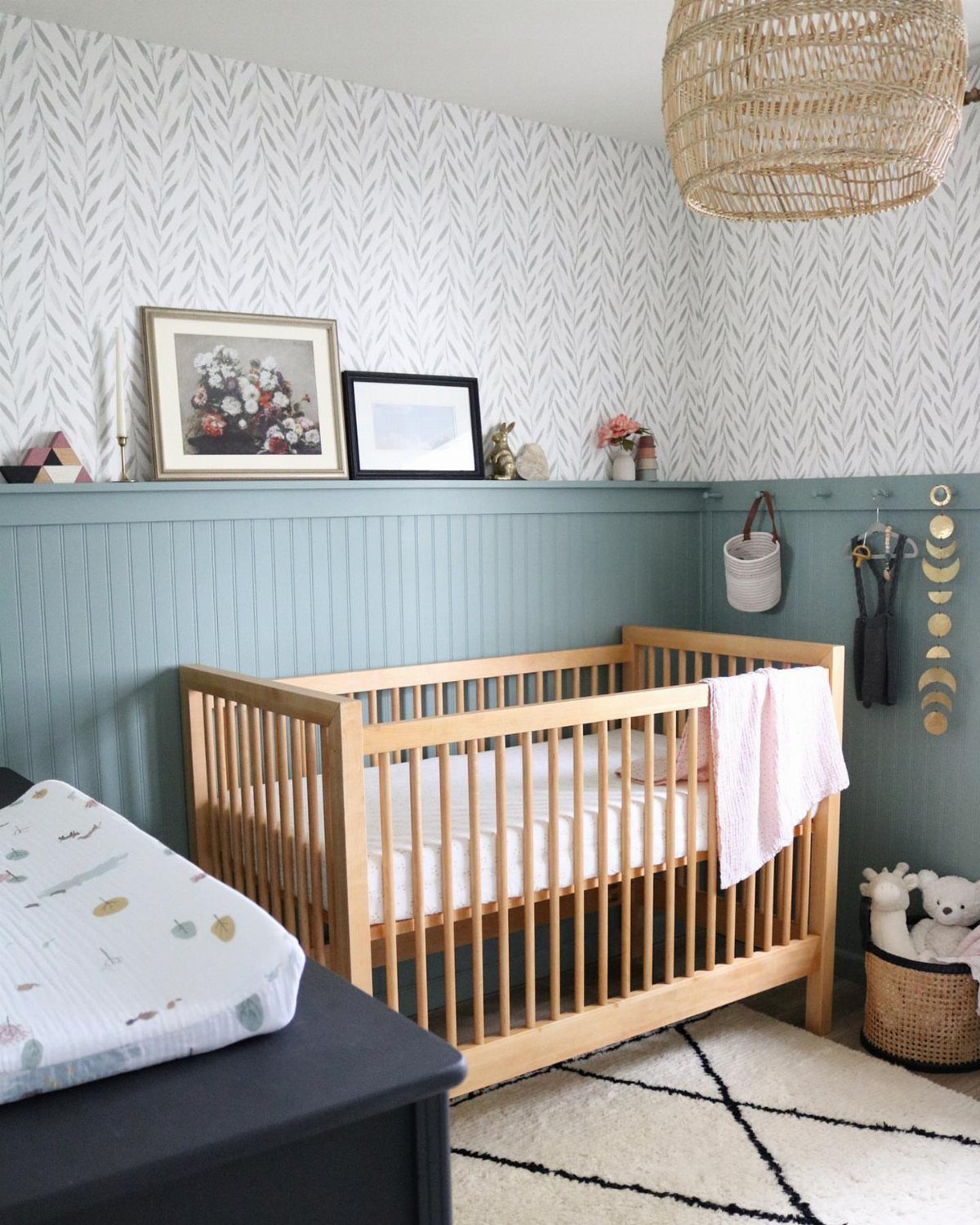 Wooden crib in a nursery with blue-gray wainscoting and patterned wallpaper walls.