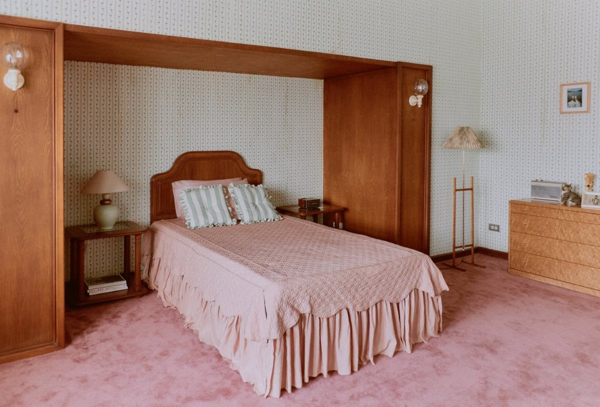 Single bed with pink ruffled bedding in a room with pink carpet.