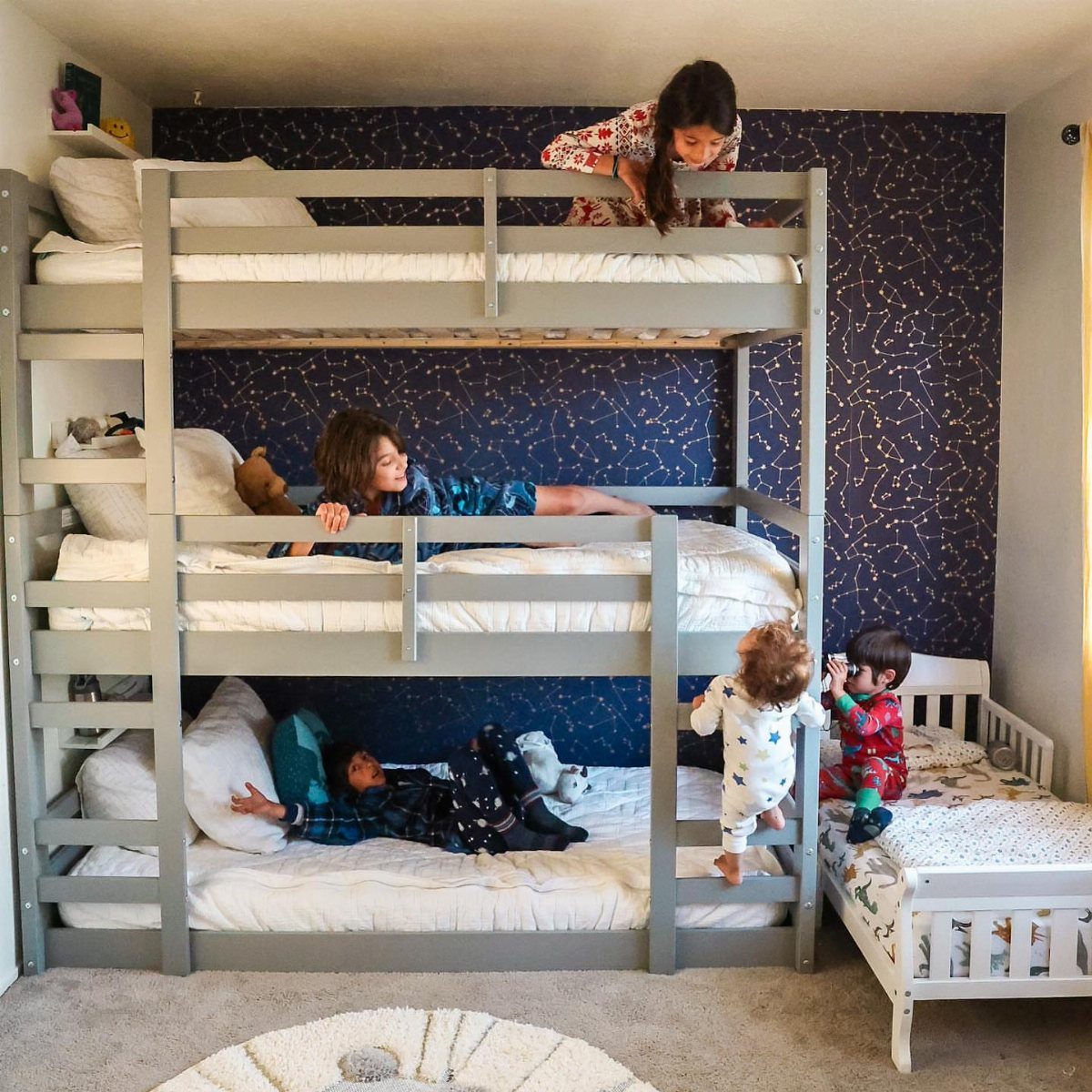 Triple-decker grey bunk bed with children and a small white toddler bed nearby.