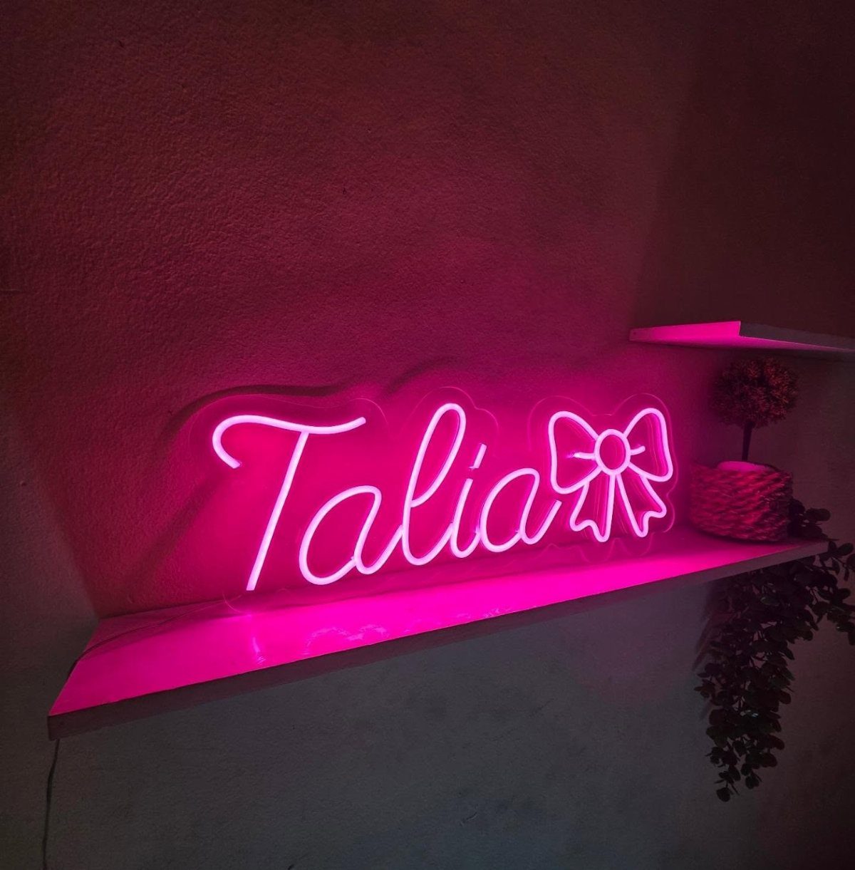Pink neon LED sign spelling Talia with a bow on a white shelf