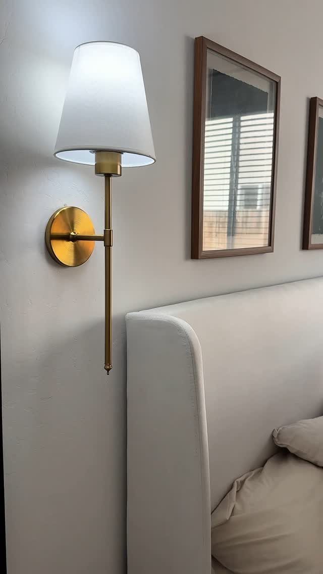 Gold wall sconce with white shade mounted beside a white upholstered bed headboard.