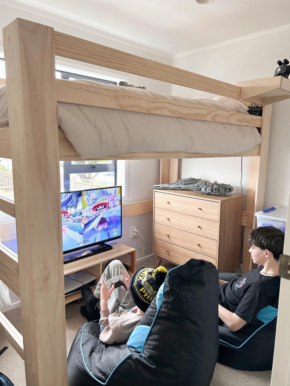 Wooden loft bed with a gaming area and bean bags underneath in a bedroom.