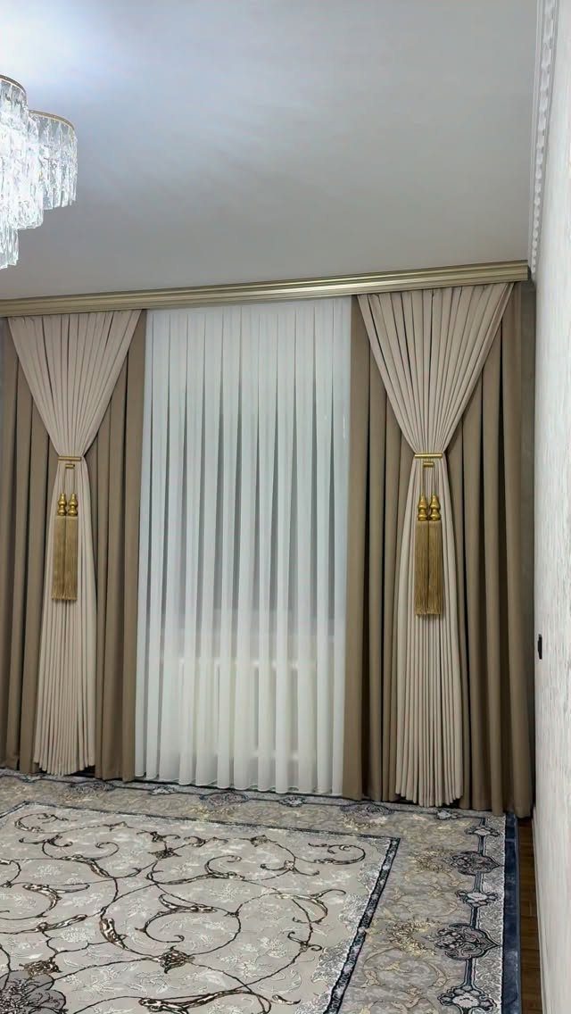 Beige and white flowing drapes with gold tassels over a patterned grey rug.