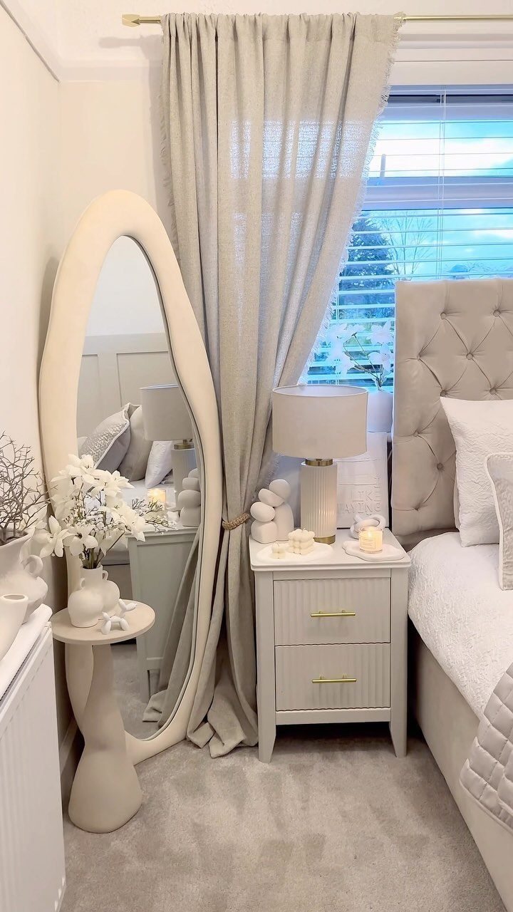 Small luxury bedroom with neutral tones, oval mirror, and tufted grey headboard.