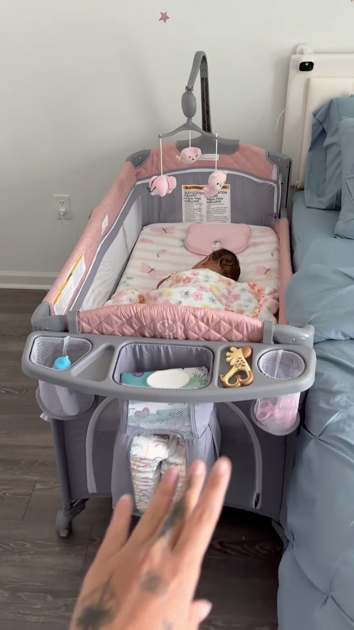 Baby sleeping in a pink and grey mini crib next to a bed