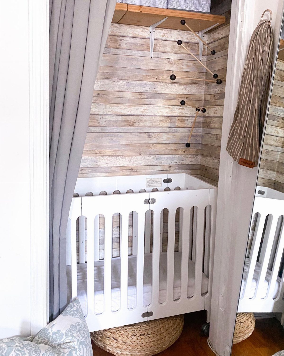 White mini crib tucked into a closet nursery with wood-paneled walls and grey curtains.