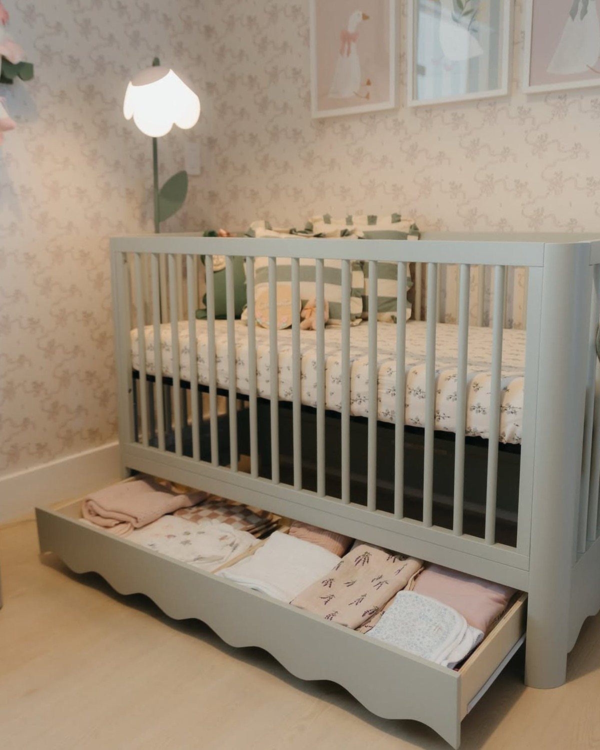 Light grey mini crib with an open storage drawer containing folded baby clothes.