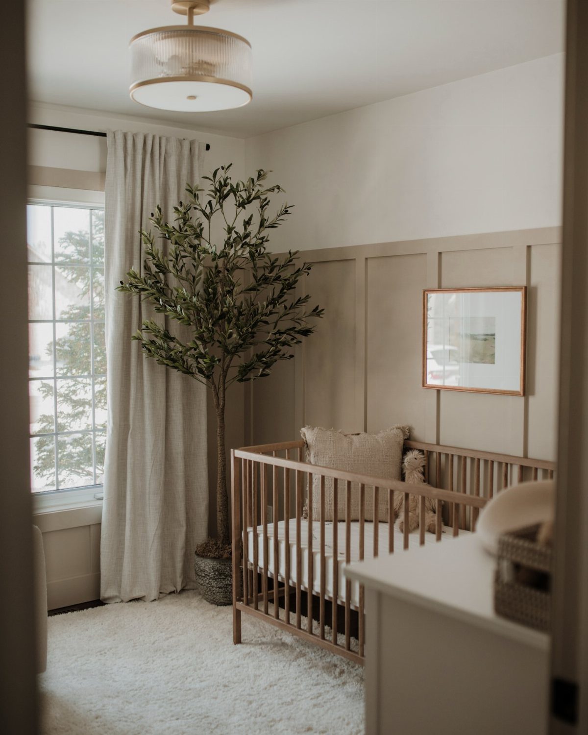 Wooden crib in a neutral nursery with beige wall paneling and olive tree.
