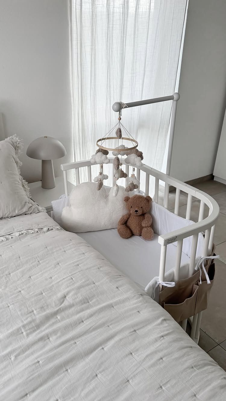White baby crib with teddy bear placed next to a bed in small room