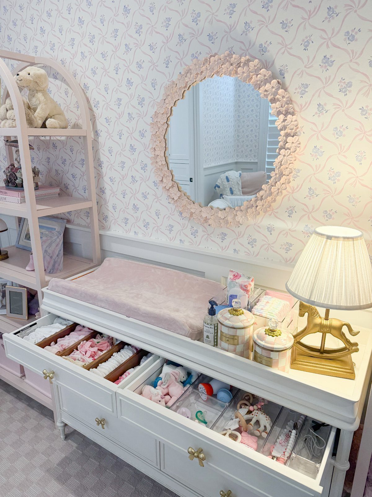 White dresser changing table with open drawers and a round floral mirror above.