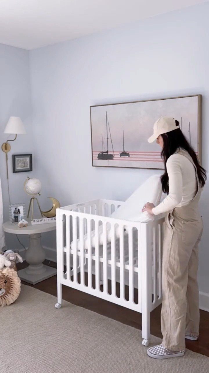 Woman adjusting bedding in a white mini crib within a light blue nursery.