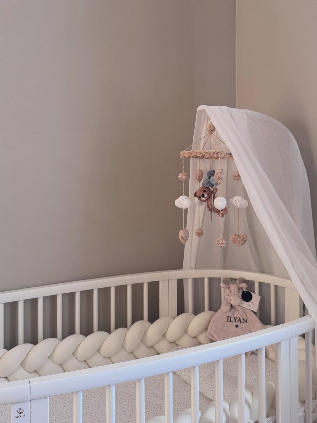 White oval baby crib with a braided bumper and a hanging bear mobile.