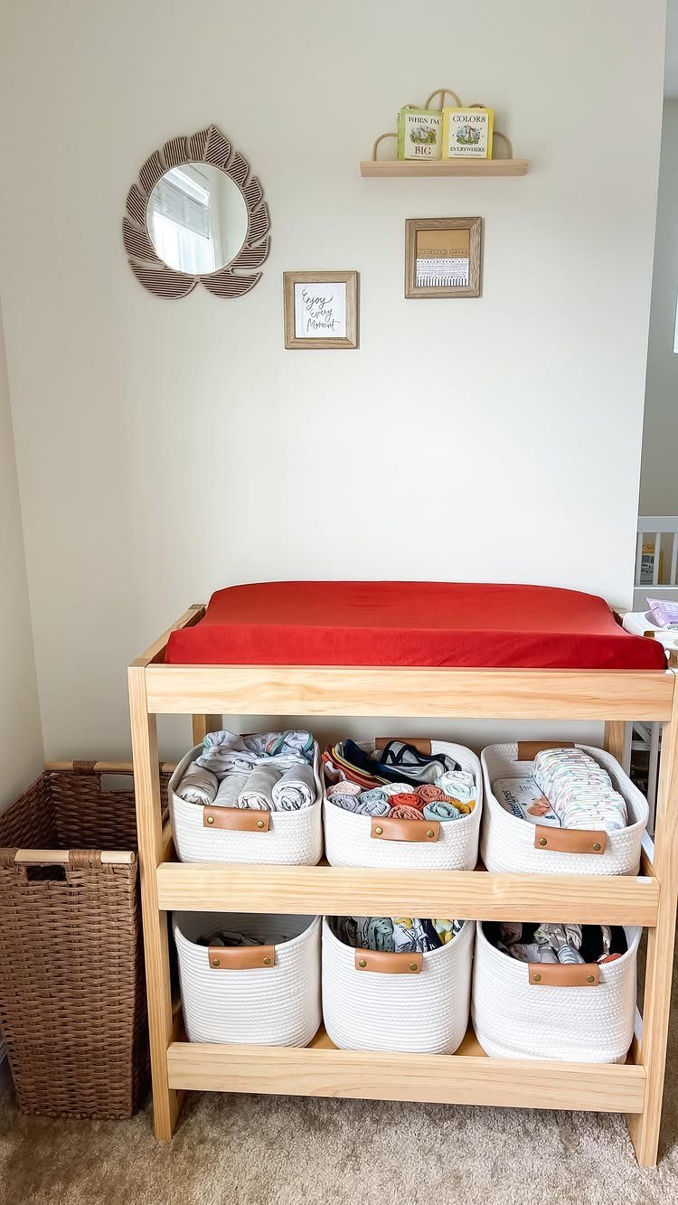 Wooden changing table with red pad and white storage bins in a nursery.