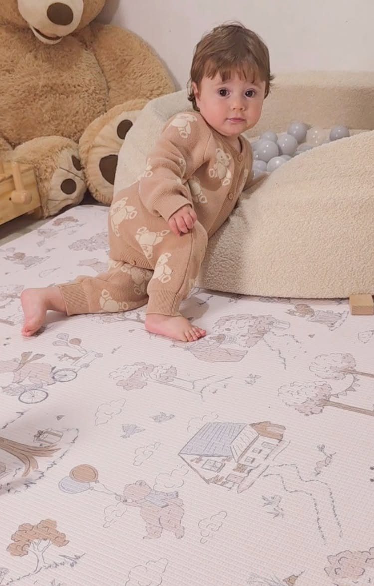 Baby in tan outfit sitting on a patterned playmat next to a ball pit.