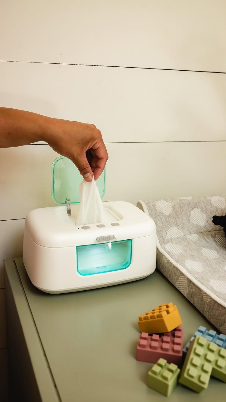 Hand pulling a baby wipe from a white electric wipe warmer on a dresser.