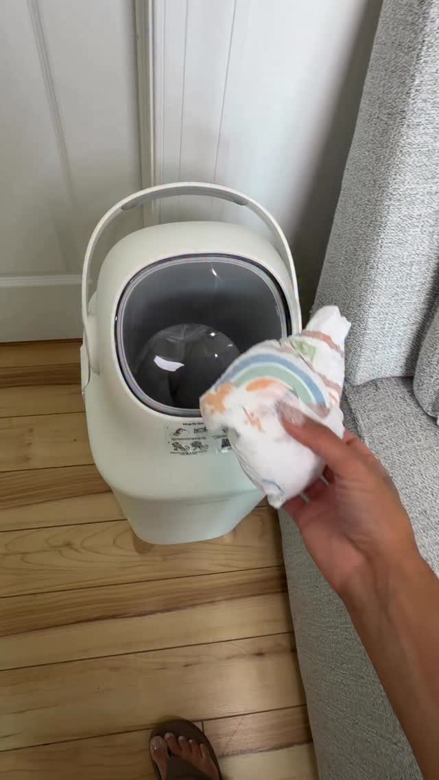White diaper pail on a wooden floor with a hand holding a diaper.